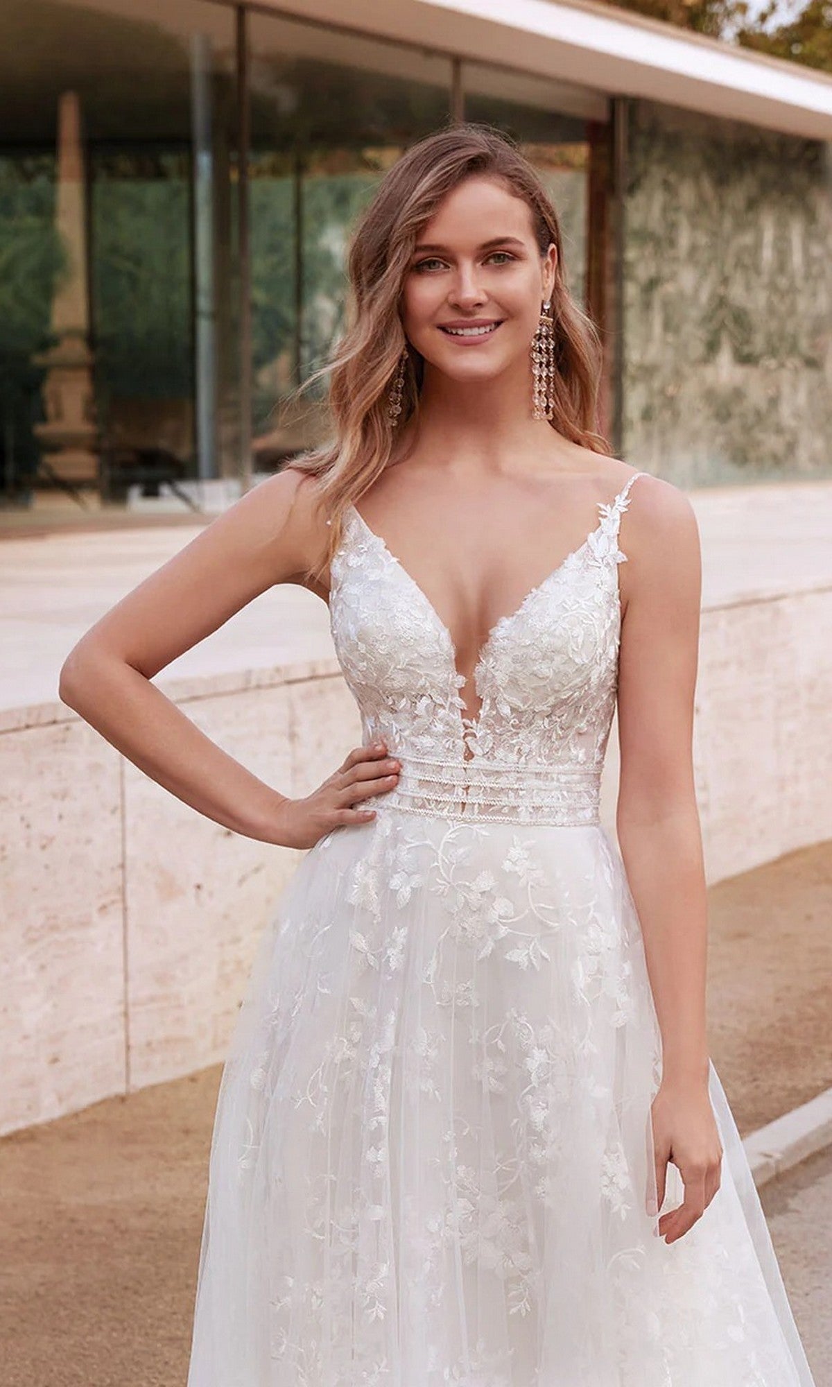 A woman stands outdoors, smiling at the camera in the Alyce 7036 Long White Dress with a deep V neckline and delicate floral appliqués.